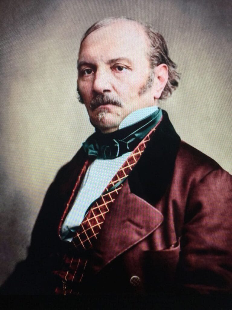 etrato de um homem com expressão séria, vestindo paletó vinho, colete com padrão geométrico e gravata verde, sobre fundo neutro