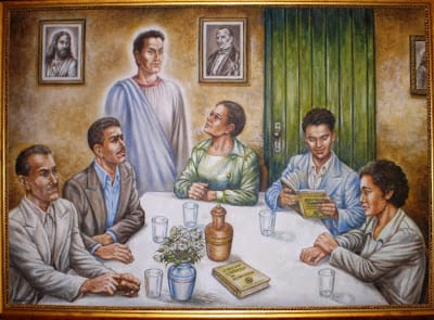 Uma pintura retrata seis homens sentados em torno de uma mesa branca, envolvidos em uma reunião ou estudo. Cinco deles estão sentados, e um homem, vestido com uma túnica branca com faixa azul, está em pé, olhando para os outros. Os homens apresentam expressões de atenção e interesse. Sobre a mesa há vários copos de vidro, um vaso com flores, um jarro de cerâmica e um livro intitulado 'O Livro dos Médiuns'. Ao fundo, duas molduras com retratos: uma figura que lembra Jesus Cristo e outra de um homem de aparência histórica. A parede de fundo é bege e há uma porta verde à direita. A pintura transmite um ambiente de estudo, reflexão e espiritualidade.
