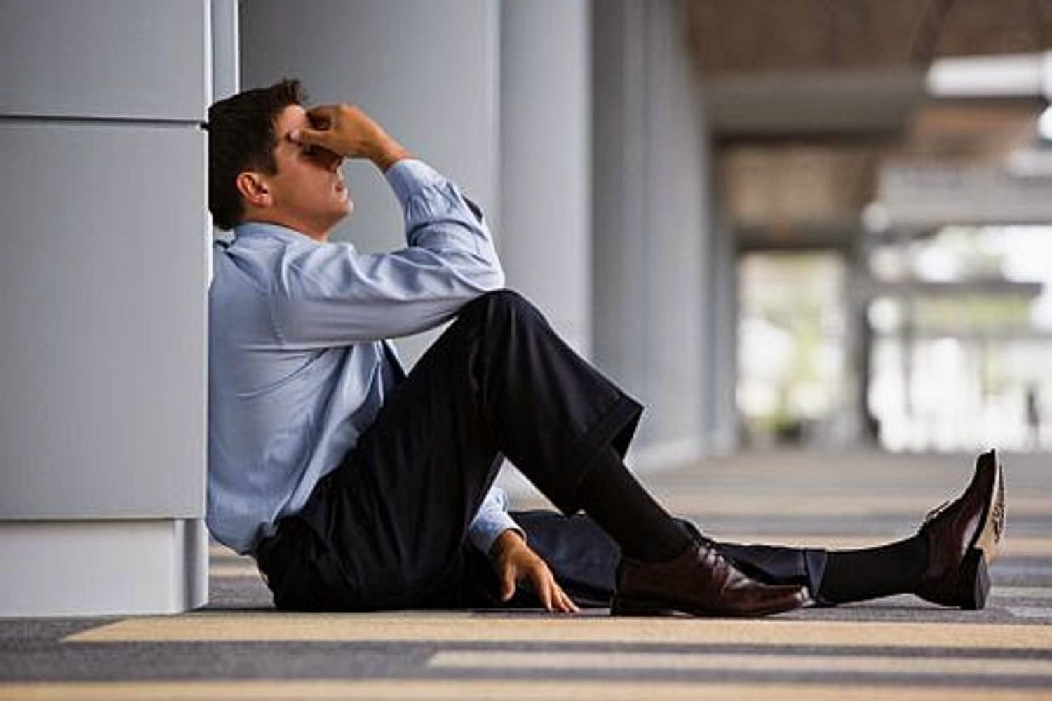 Homem de terno sentado no chão, encostado em uma parede, segurando o rosto em sinal de arrependimento e angústia.
